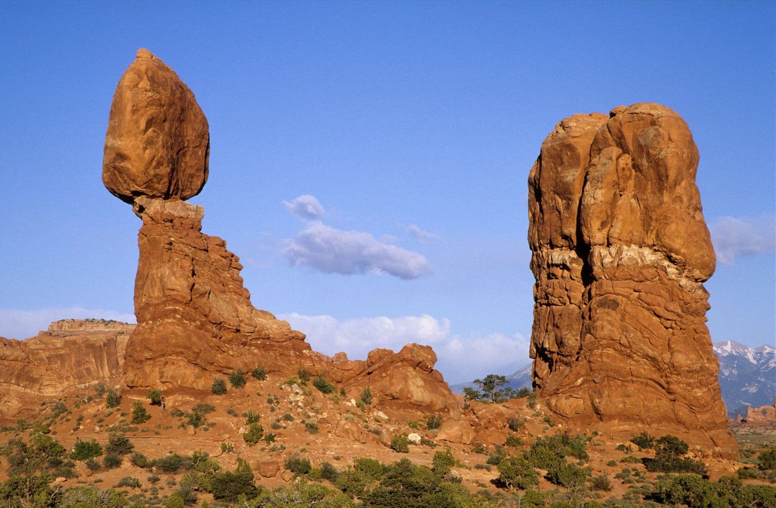 Balanced Rock sandstone formation rises above desert brush with snow-capped La Sal Mountains visible in Arches National Park.
