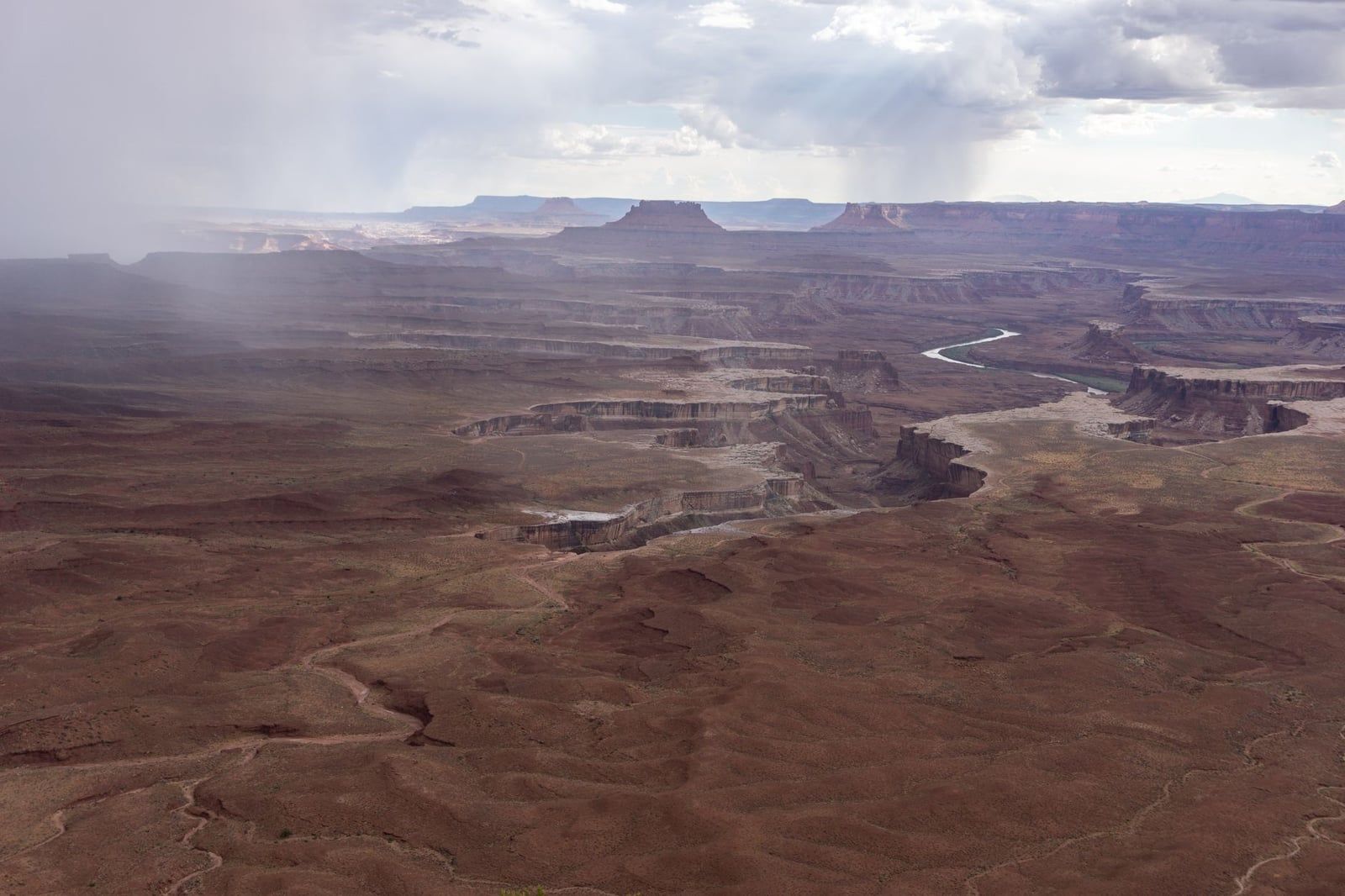 A wide view from the Green River Overlook in Canyonlands National Park, showing the Green River winding through deep sandstone canyons under rain clouds.